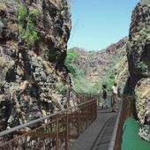 2 people walking along a metal walkway with a steel rail fence on both sides, which runs between 2 tall stone cliffs