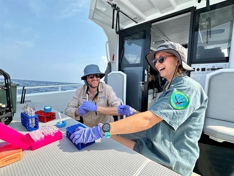 Two people in green shirts and widebrim hats sitting on the back of a boat at a desk covered in medical sample vials.
