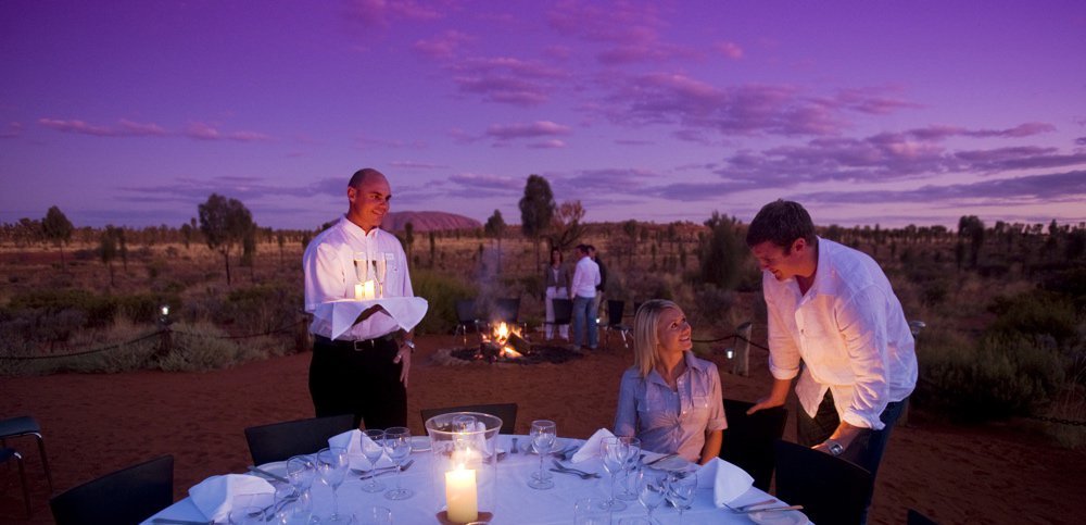 Woman seated at an outdoor table during dusk with Uluru in the backgrounds She is talking to a man as a waiter holds two glasses of sparkling wine.